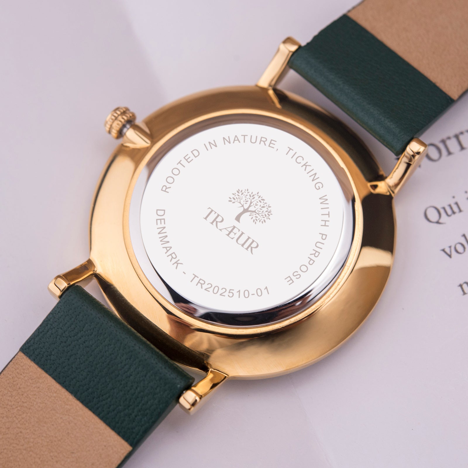 Gold watch back with engraved tree logo and green leather strap on white surface