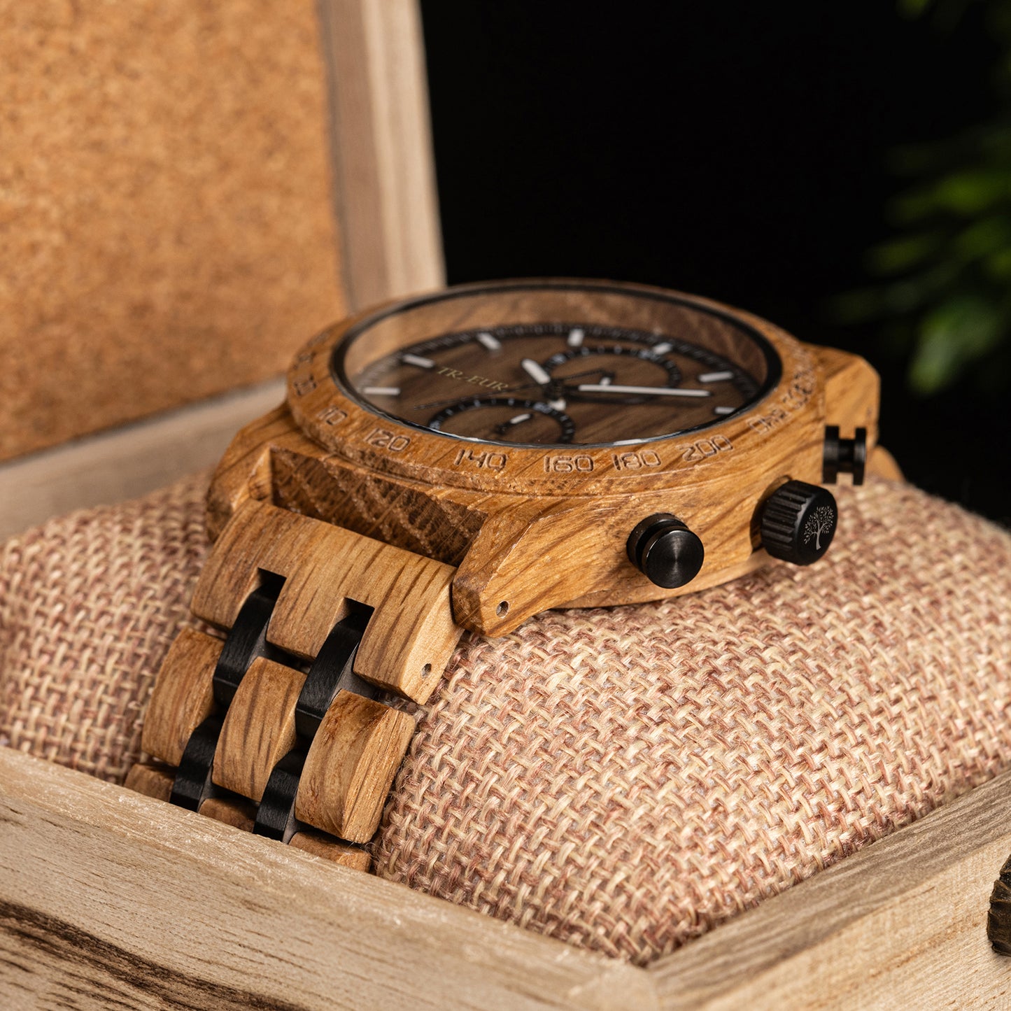 Close-up of wooden wristwatch with black dials and chronograph, resting on textured cushion in wooden box
