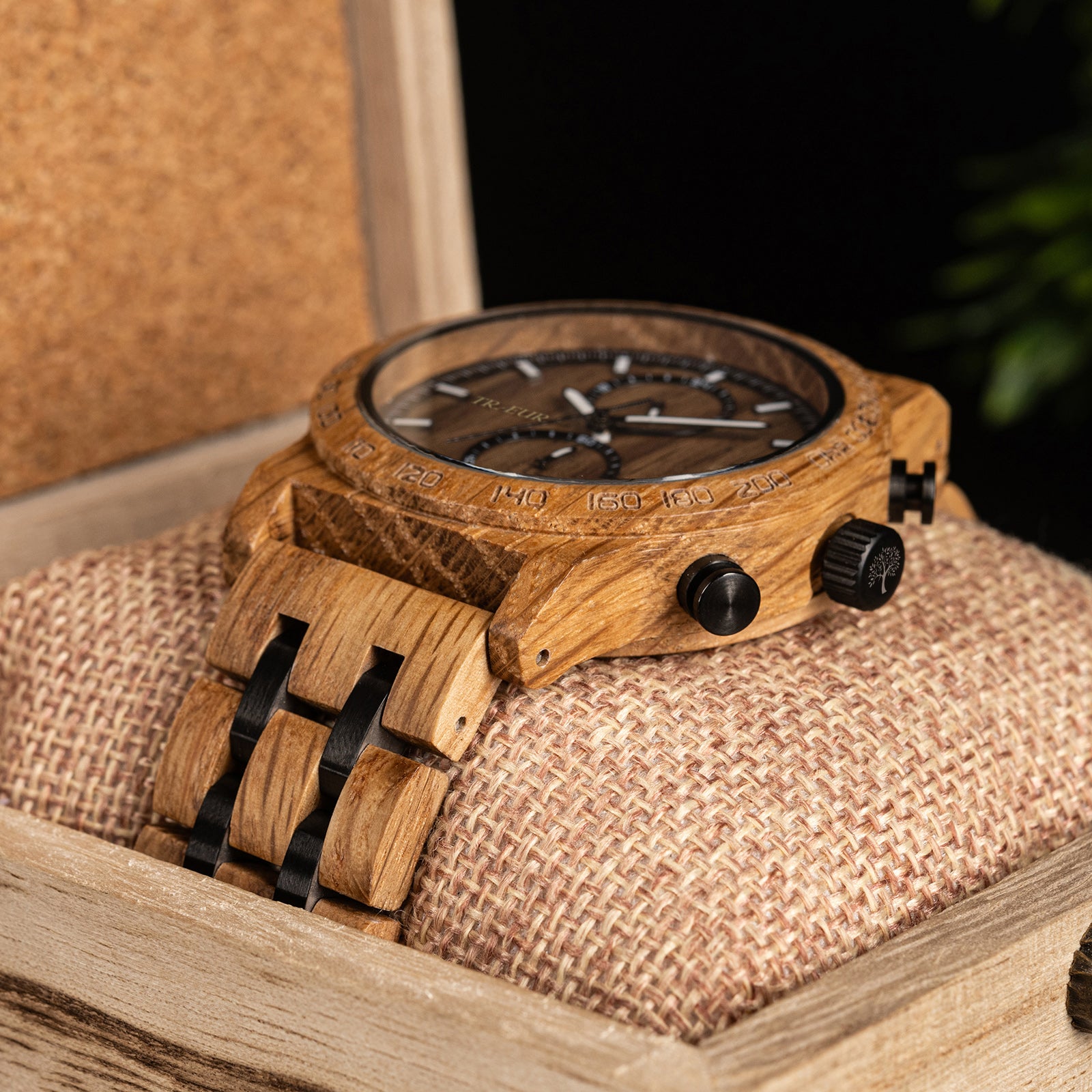 Close-up of wooden wristwatch with black dials and chronograph, resting on textured cushion in wooden box