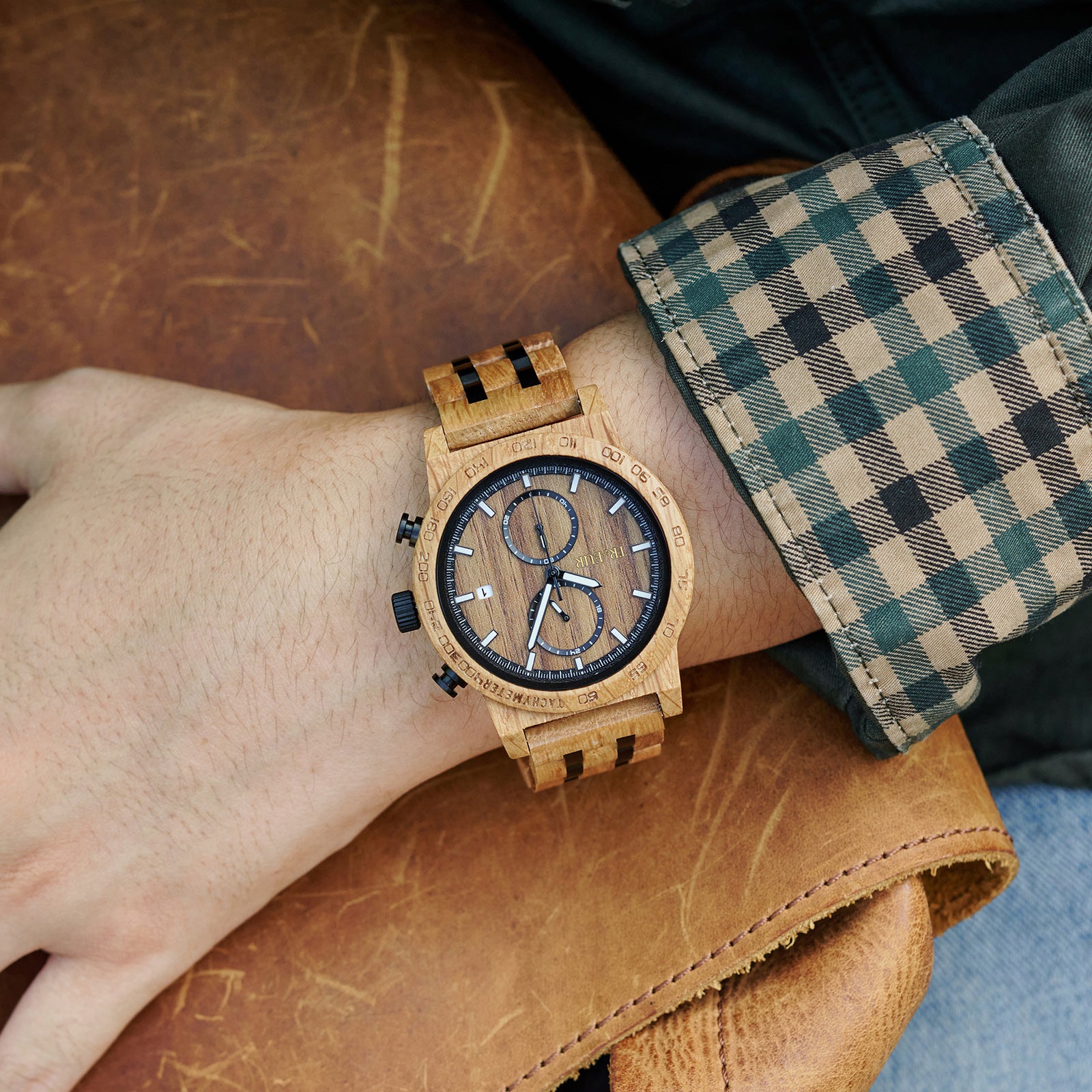 Close-up of wrist wearing wooden chronograph watch with black accents and checkered shirt cuff on leather surface